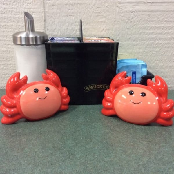 Two smiling red crab figurines flank a sugar dispenser and a Smucker's caddy on a green table.