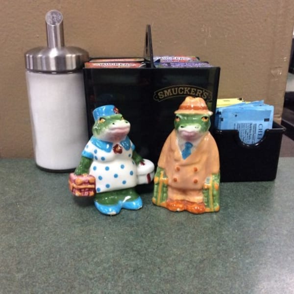 Two frog figurines, a sugar dispenser, and a Smucker's caddy on a cafe table.