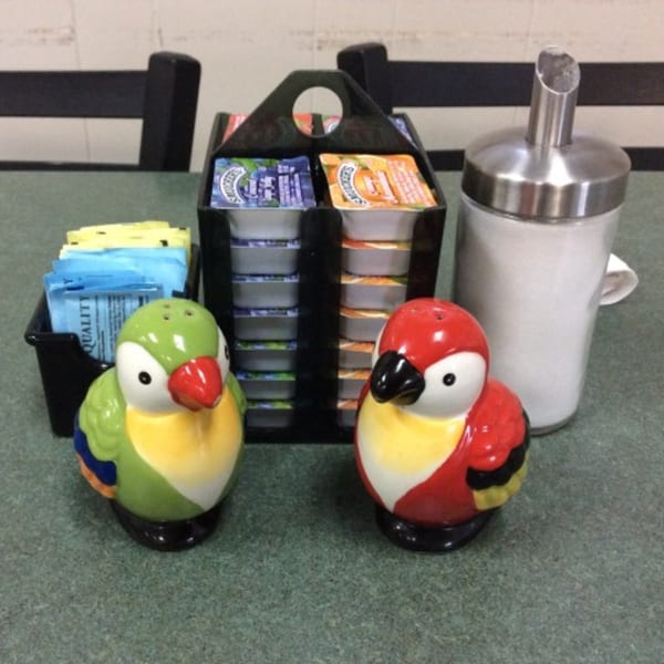 Two colorful parrot-shaped salt and pepper shakers on a table with condiments and sugar.