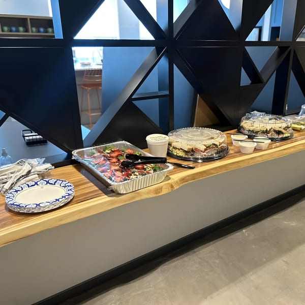 A buffet line with a strawberry salad, sandwiches, paper plates, and plastic cutlery on a wooden counter.