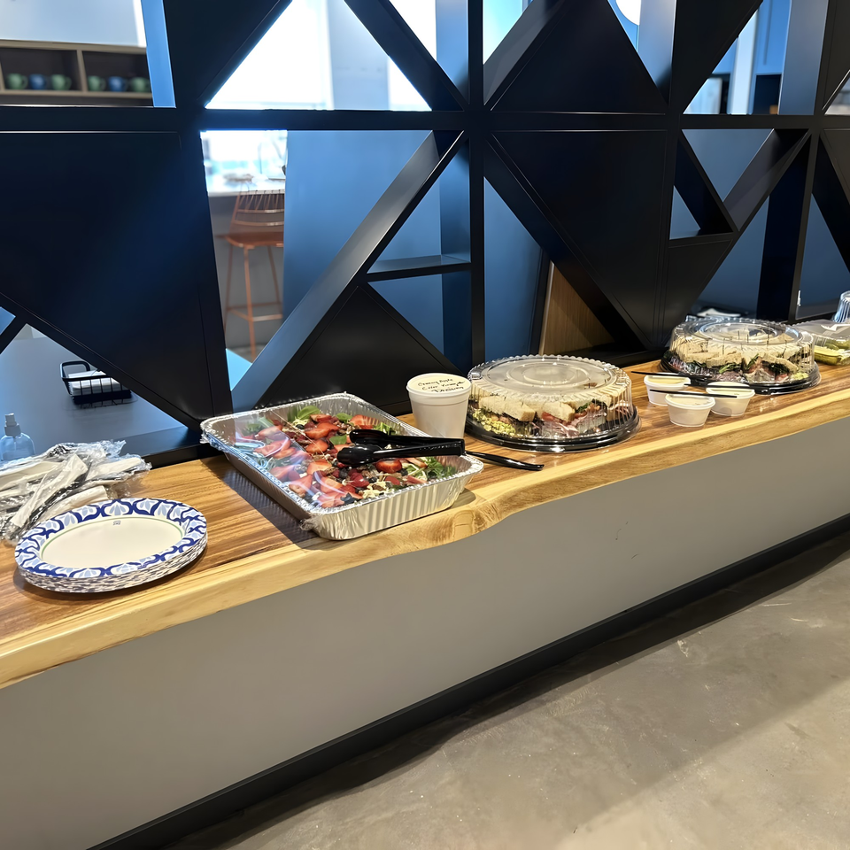 A buffet line with a strawberry salad, sandwiches, paper plates, and plastic cutlery on a wooden counter.