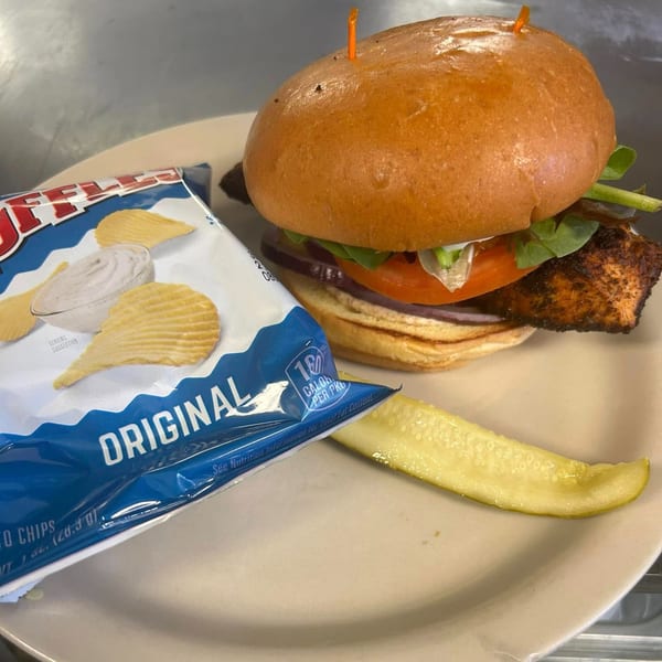 Shakers American Café's blackened salmon sandwich with lettuce, tomato, onion, a pickle, and chips.