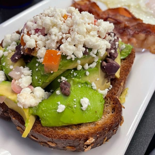 Avocado toast with feta, diced tomatoes, and olives, served with bacon and eggs.