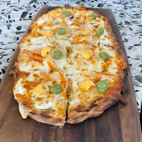 A rectangular flatbread pizza with melted cheese, orange toppings, and green and yellow dollops on a wooden board.