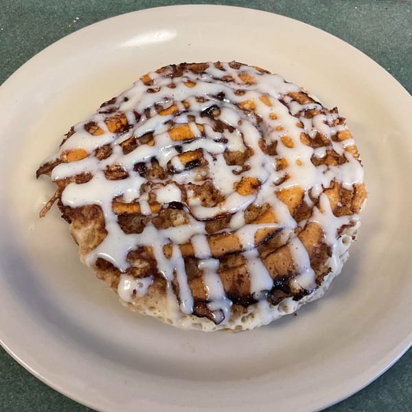 A large, round cinnamon roll with white icing drizzled over a golden-brown pastry on a white plate.
